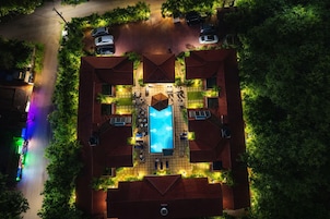 An aerial view of a property with a swimming pool surrounded by lounge chairs and umbrellas, a covered patio area, and a street with parked cars.