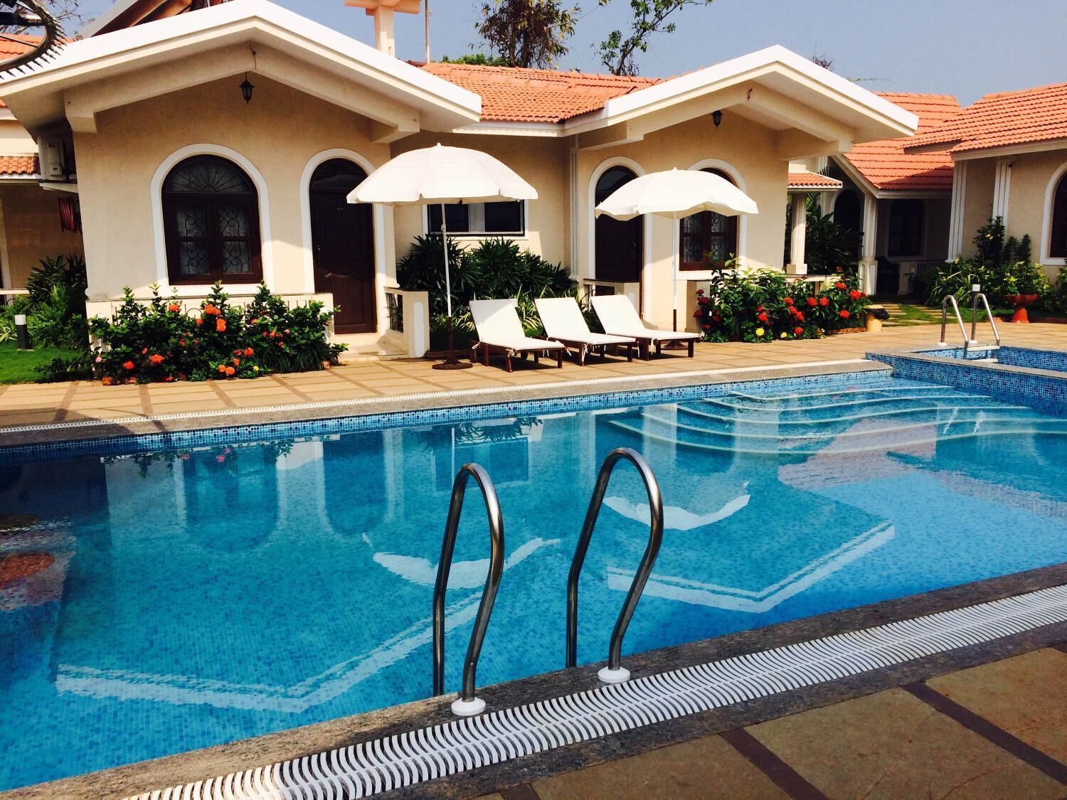 A swimming pool with a tiled edge, surrounded by lounge chairs and umbrellas, with a building featuring arched windows and a tiled roof in the background.