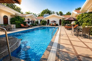 A swimming pool with lounge chairs and umbrellas, surrounded by greenery and buildings with a tiled roof.