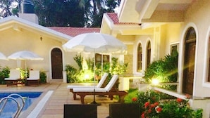 An outdoor pool area with lounge chairs and umbrellas, surrounded by greenery and a well-lit patio. - Jacks Resort (Vagator)