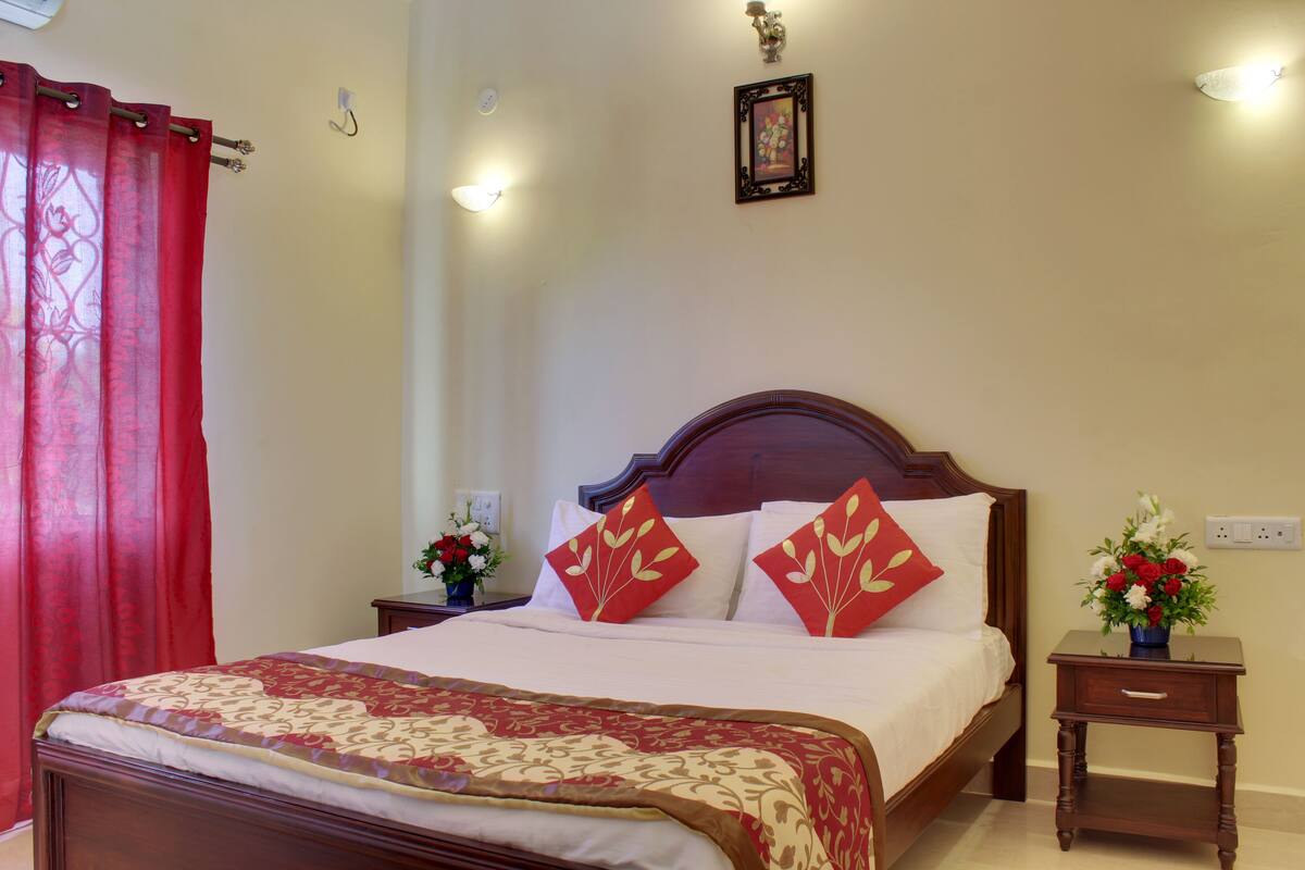 A neatly made bed with a red and gold patterned bedspread, two nightstands with flower vases, and a wall-mounted picture.