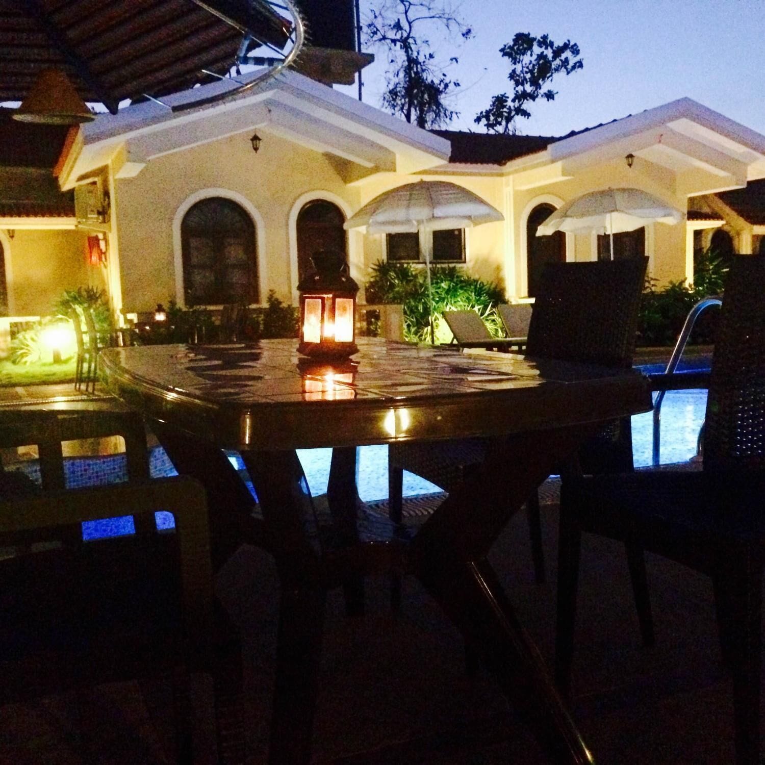 An outdoor pool area with a round table and chairs, surrounded by a building with arched windows and a tiled roof.