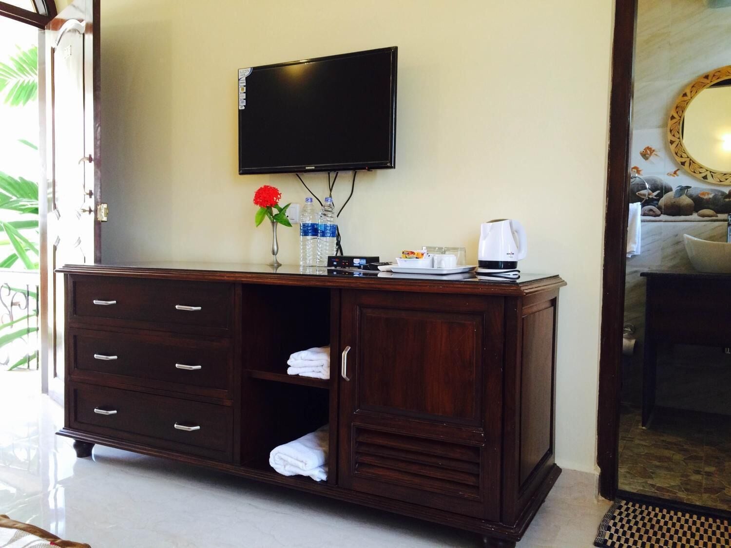 Une chambre d’hôtel avec un meuble en bois, un téléviseur à écran plat, un vase avec une fleur rouge, une machine à café et un miroir doté d’un cadre décoratif.