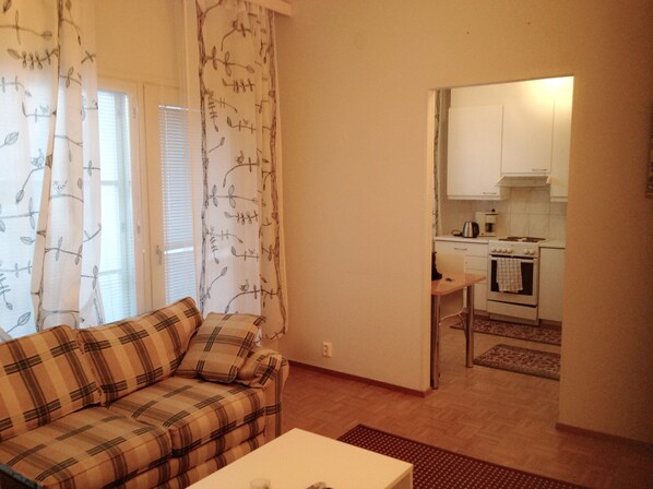 A living room with a plaid sofa and a small table, leading to a kitchen with white cabinets and a stove. - City Apartment Lappeenranta (Lappeenranta)