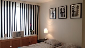 A bedroom with a bed, a bedside table, a lamp, a dresser, and a window with vertical blinds. - City Apartment Lappeenranta (Lappeenranta)