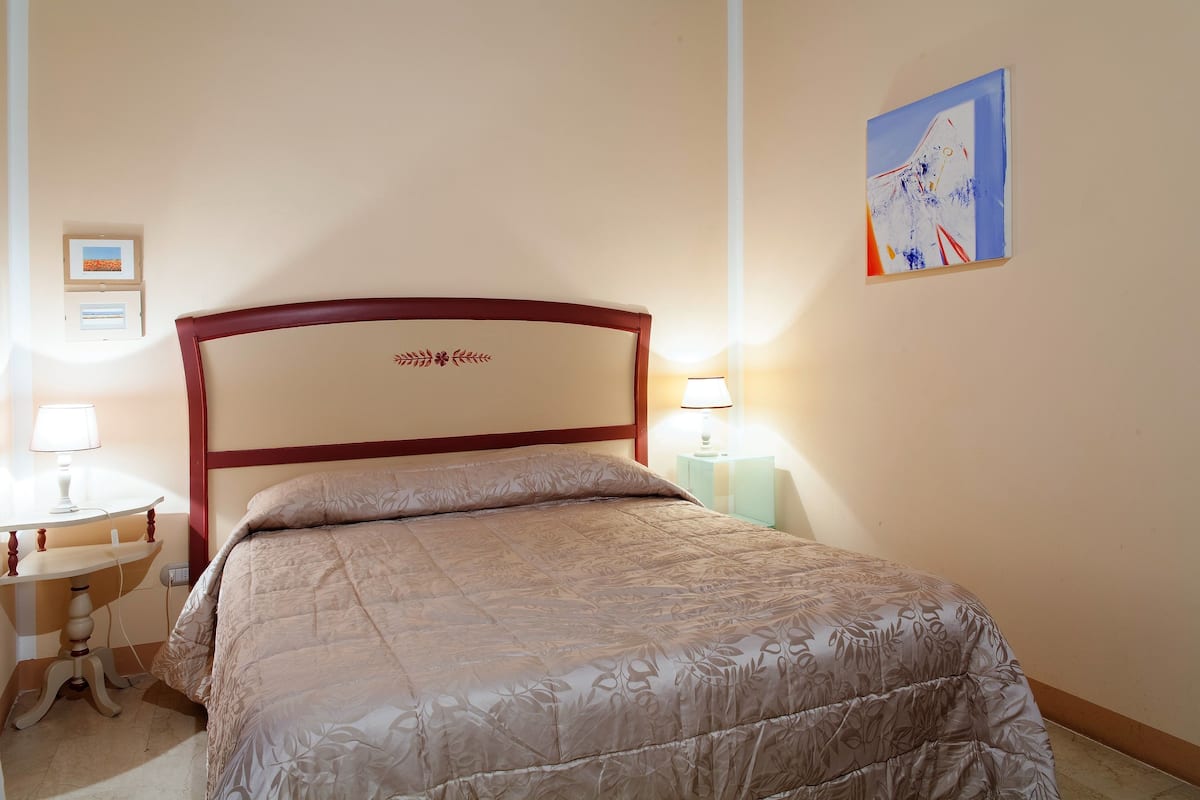 A neatly made bed with a floral-patterned bedspread, a wooden headboard, a bedside table with a lamp, and a wall-mounted painting.