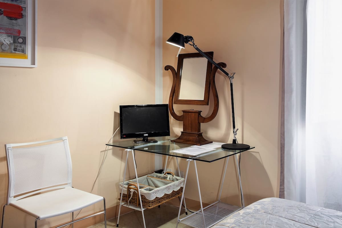 A room with a glass desk, a computer monitor, a lamp, a chair, and a bed.