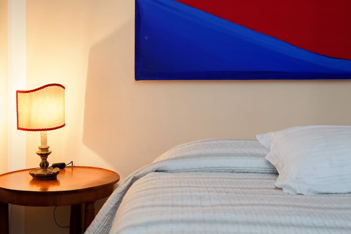 A bed with white bedding, a wooden bedside table with a lamp, and a red and blue abstract painting on the wall.