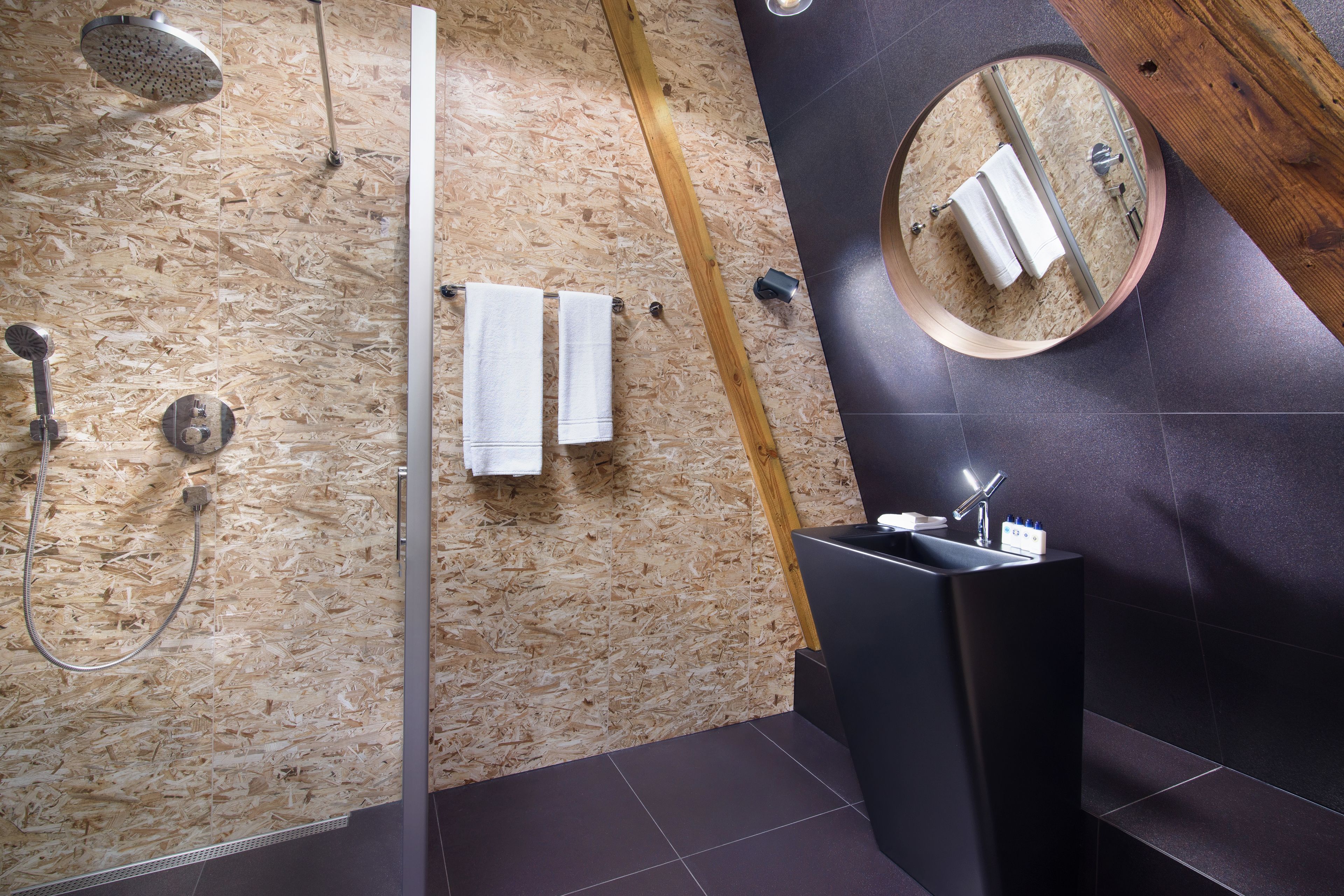 A modern bathroom with a wooden wall, a round mirror, a black sink, and a shower area.
