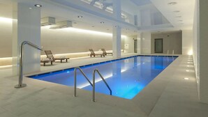 A modern indoor swimming pool with clear blue water, lounge chairs, and a well-lit ceiling.