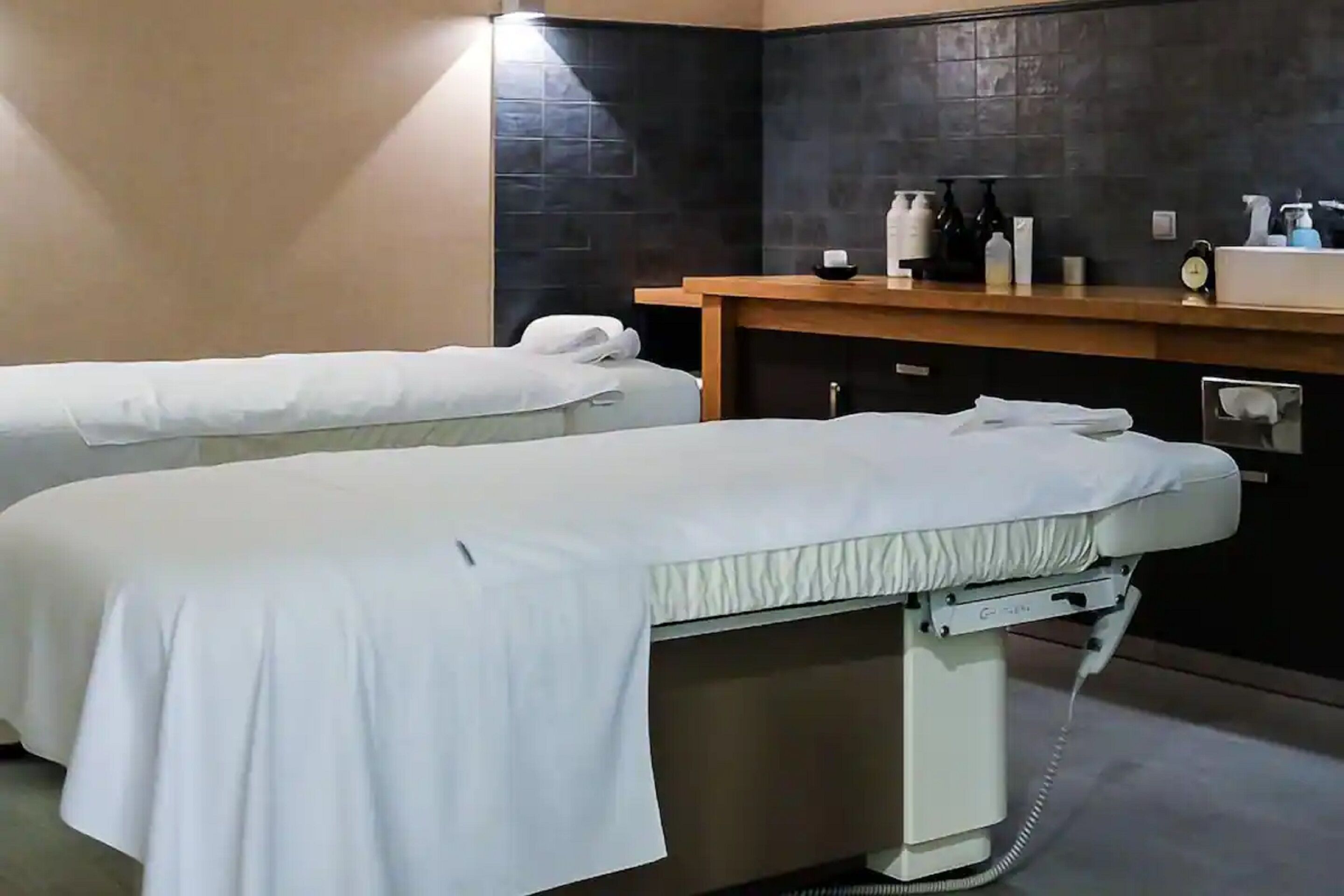 A room with two beds, a wooden counter with toiletries, and a black tiled wall.