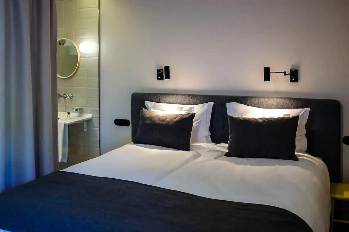 Une chambre d’hôtel avec un grand lit, deux tables de chevet, un luminaire fixé au mur et une salle de bain avec un lavabo et un miroir.