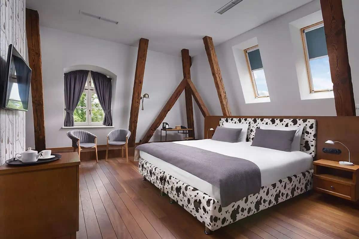 Une chambre avec un grand lit, des poutres en bois, un bureau avec une tasse et une soucoupe, deux chaises et une fenêtre avec des rideaux.