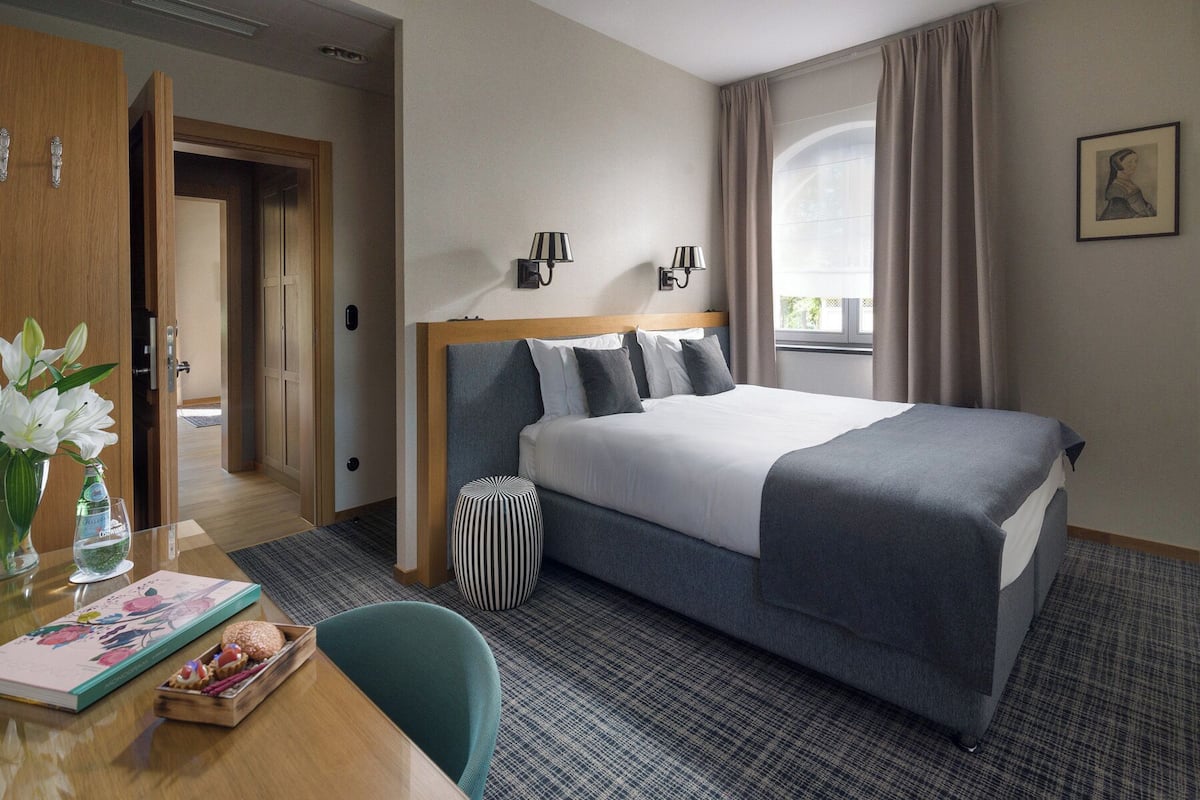 Une chambre d’hôtel avec un grand lit, un bureau avec une chaise, un vase de fleurs et une fenêtre avec des rideaux.