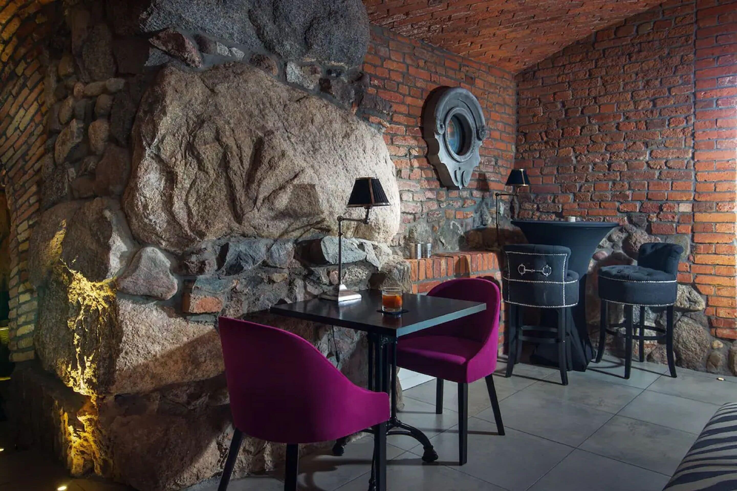 Un comedor con pared de piedra, pared de ladrillo y un espejo redondo. Hay dos sillas moradas, una mesa y dos taburetes de bar.