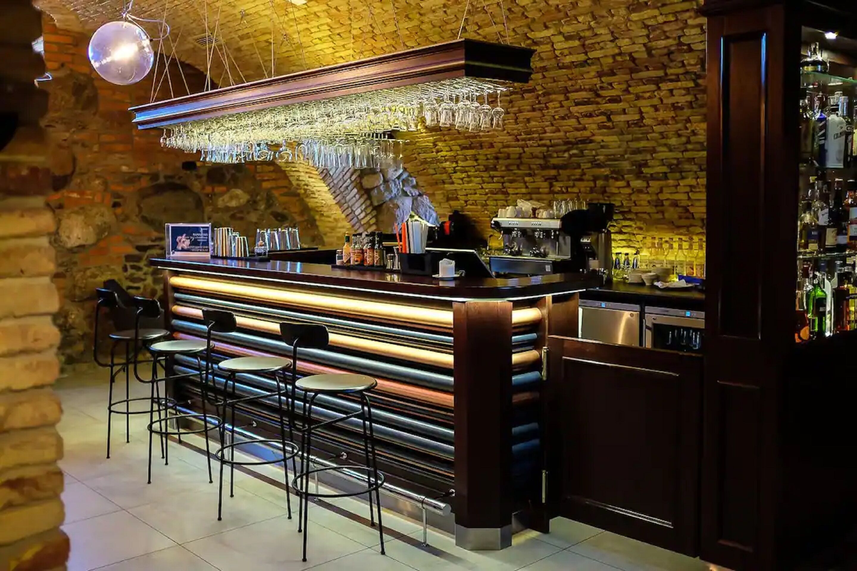 A bar area with a brick wall, bar stools, a counter with a coffee machine, and hanging glasses.