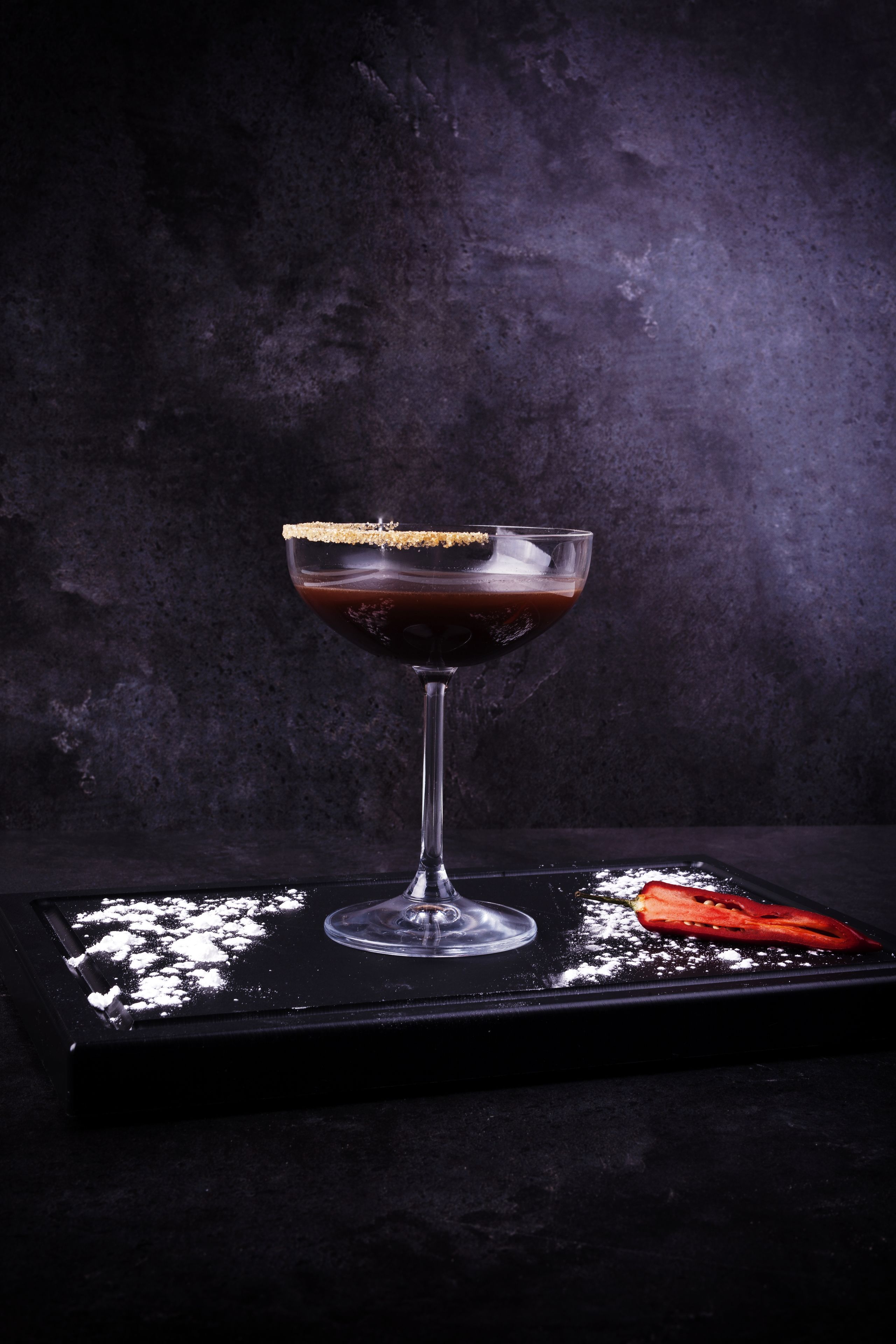 A cocktail in a glass with a salted rim, placed on a dark surface with powdered sugar and a red item beside it.