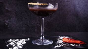 A cocktail in a glass with a salted rim, placed on a dark surface with powdered sugar and a red item beside it.