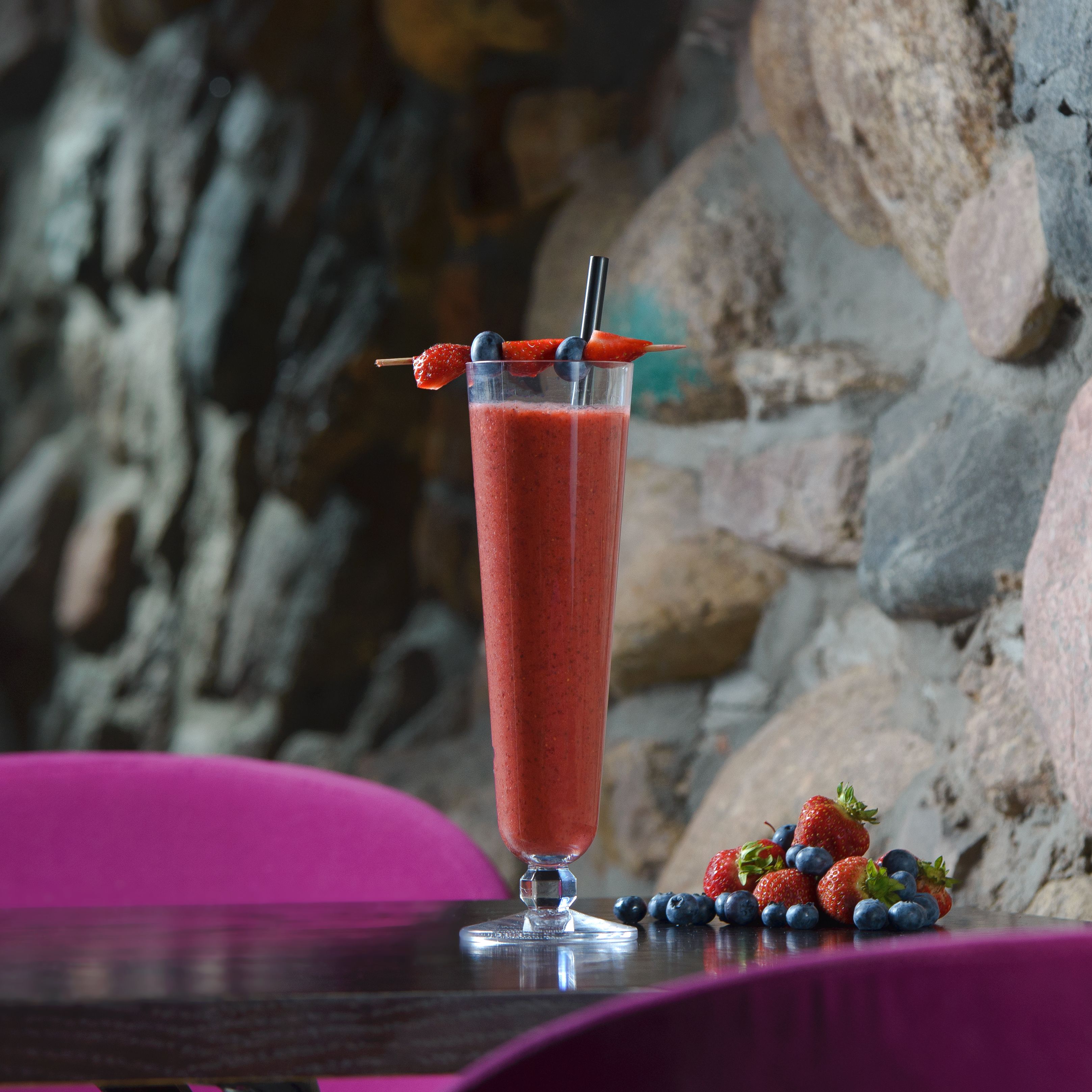 A tall glass of red beverage with a straw and fruit garnish, placed on a table with a stone wall background.