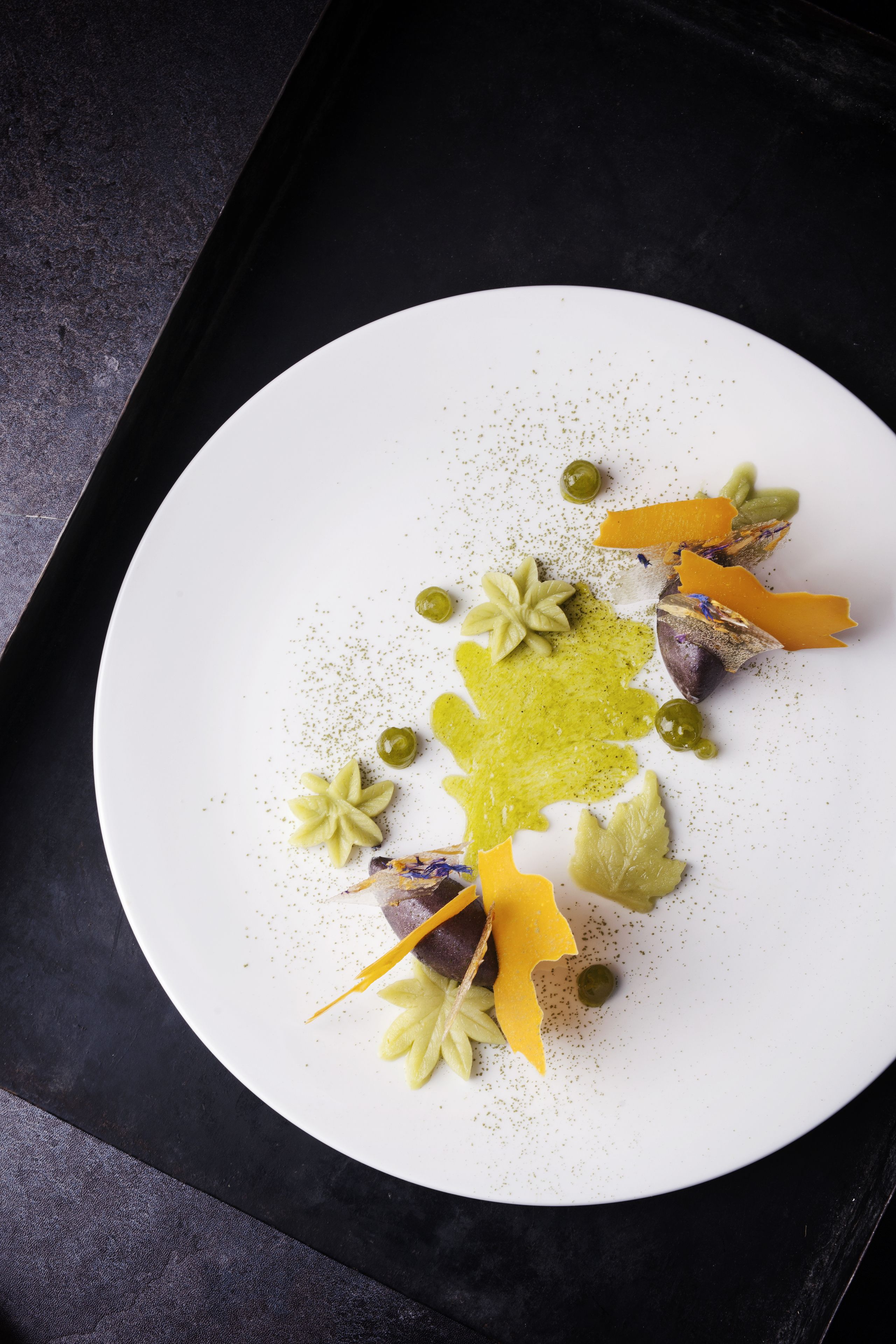 A white plate with a variety of food items, including green olives, star anise, and yellow-orange pieces, arranged on a dark surface.