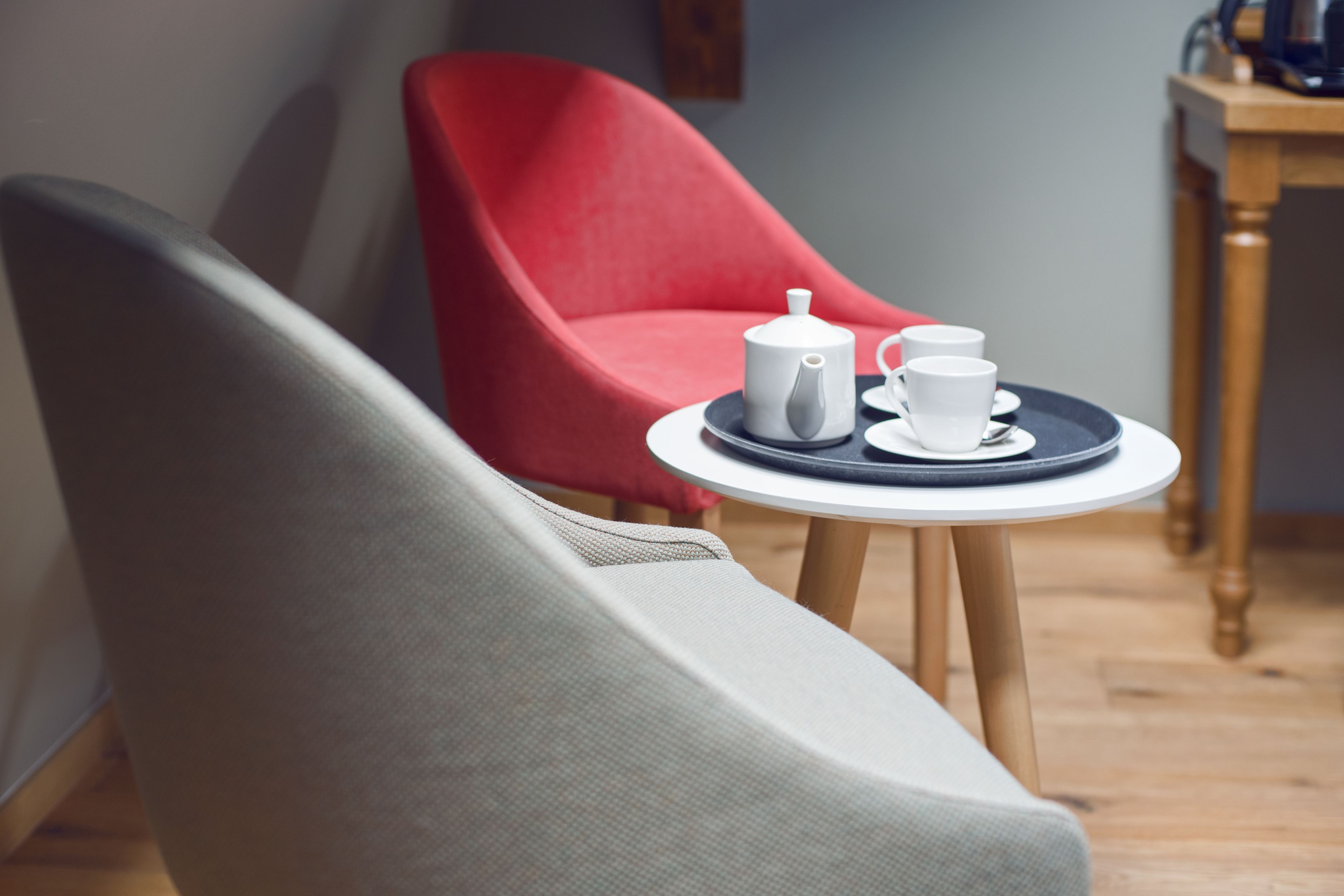 A room with a grey armchair, a red chair, and a small round table with a white teapot and cups.