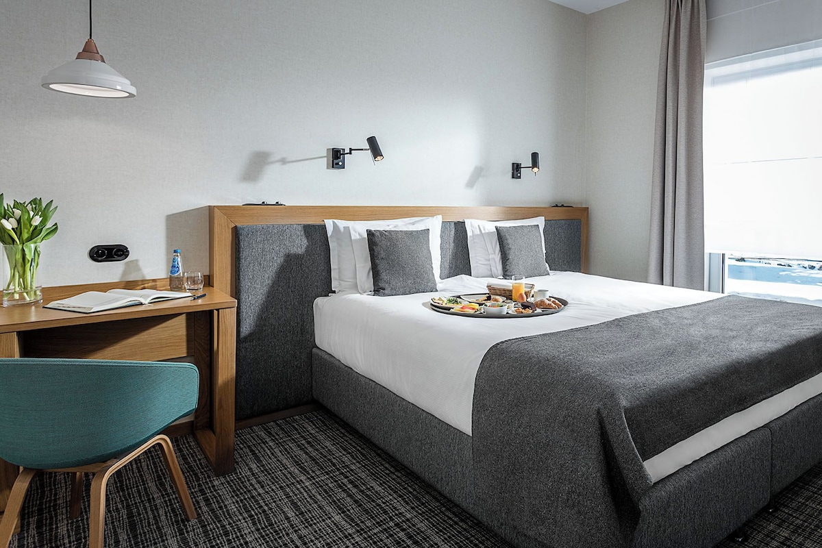 A hotel room with a bed, a desk with a chair, a window with curtains, and a view of the outdoors.