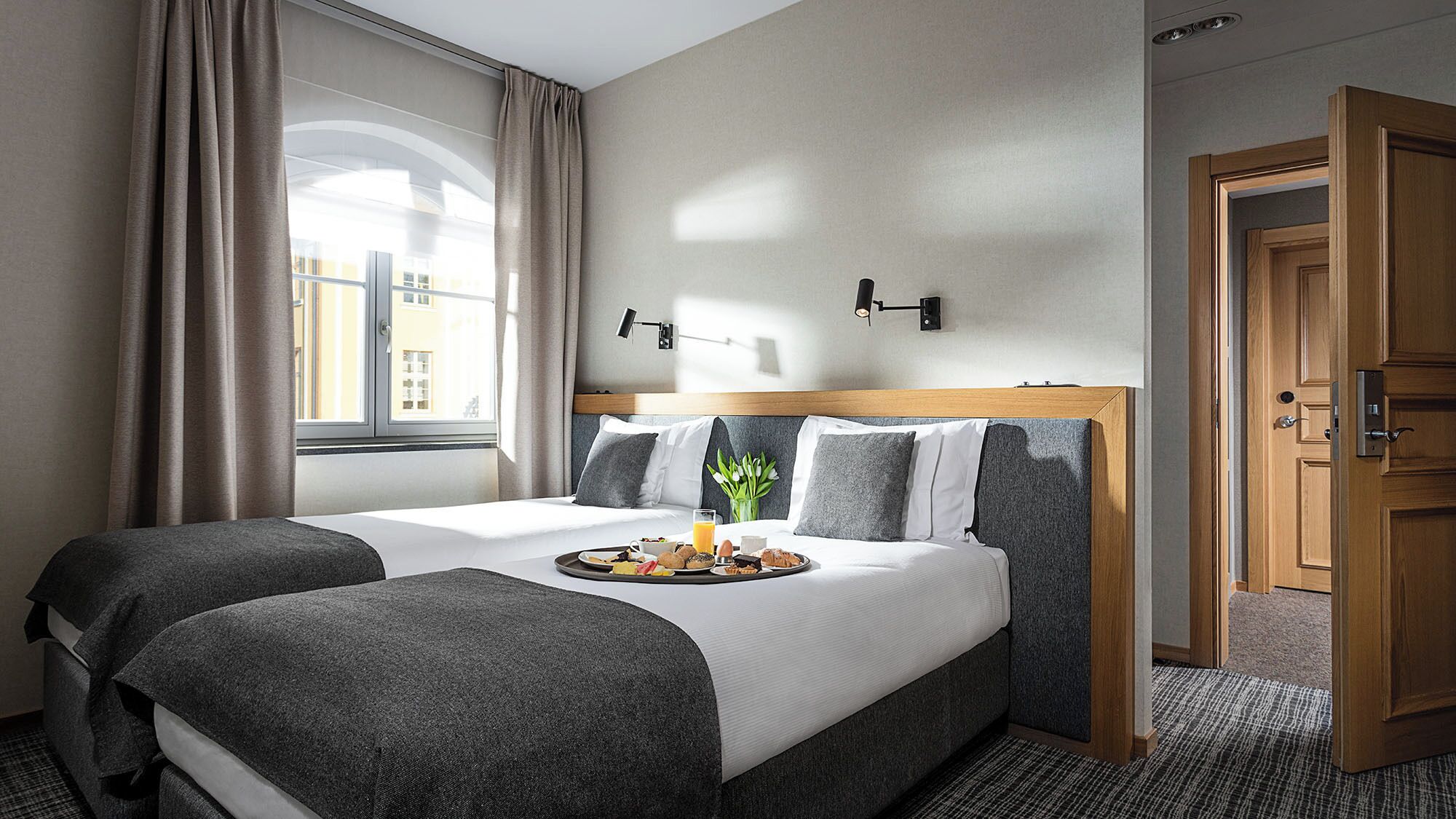 Habitación de hotel con dos camas, un carrito de desayuno, una ventana con cortinas, una lámpara de pared y una puerta.