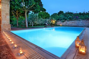 A swimming pool with a stone border, surrounded by a garden with trees and flowers, and two lit lanterns on the edge. - Vouliagmeni Villa (Vari-Voula-Vouliagmeni)