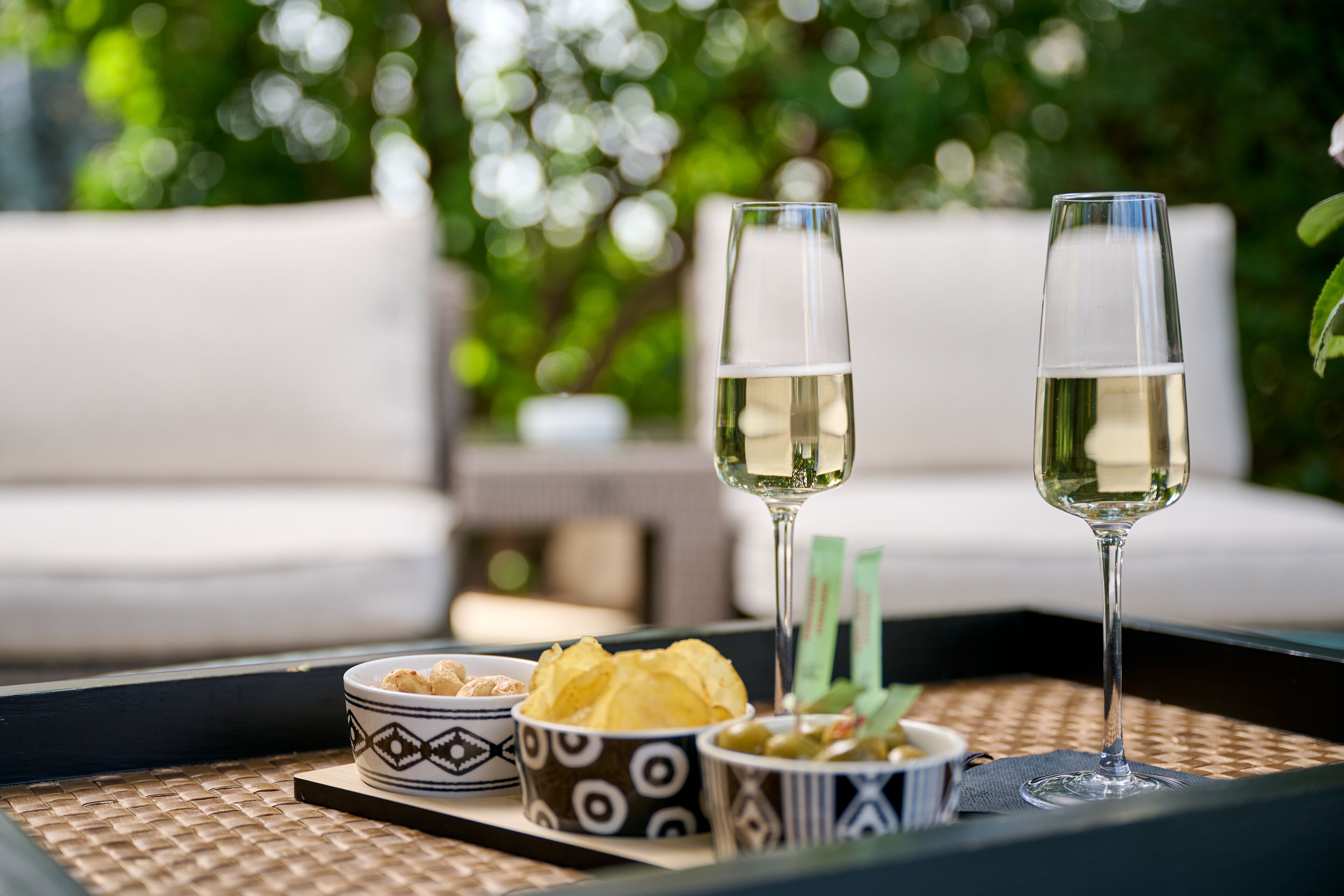 A tray with two glasses of white wine, a bowl of chips, and a bowl of olives on a woven mat.