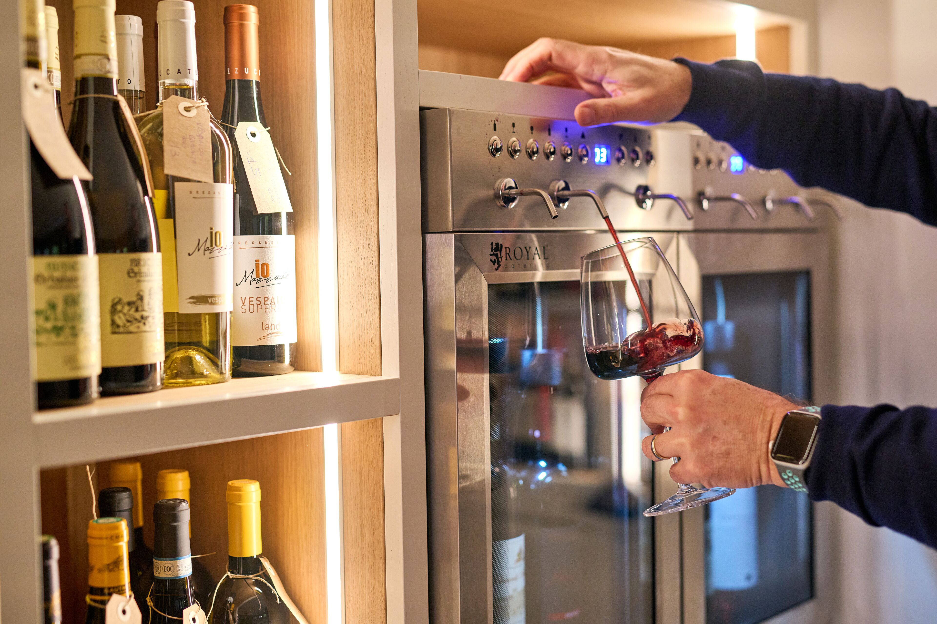En vinkøler med forskellige vinflasker og en person, der hælder vin i et glas.