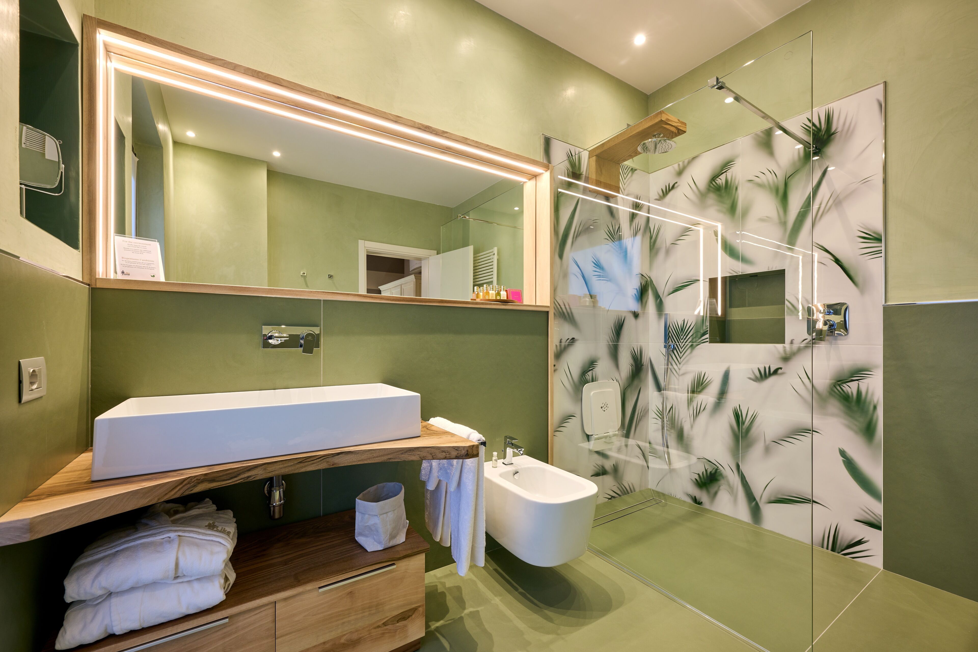 A modern bathroom with a large mirror, a sink, a bidet, and a shower with palm leaf wallpaper.