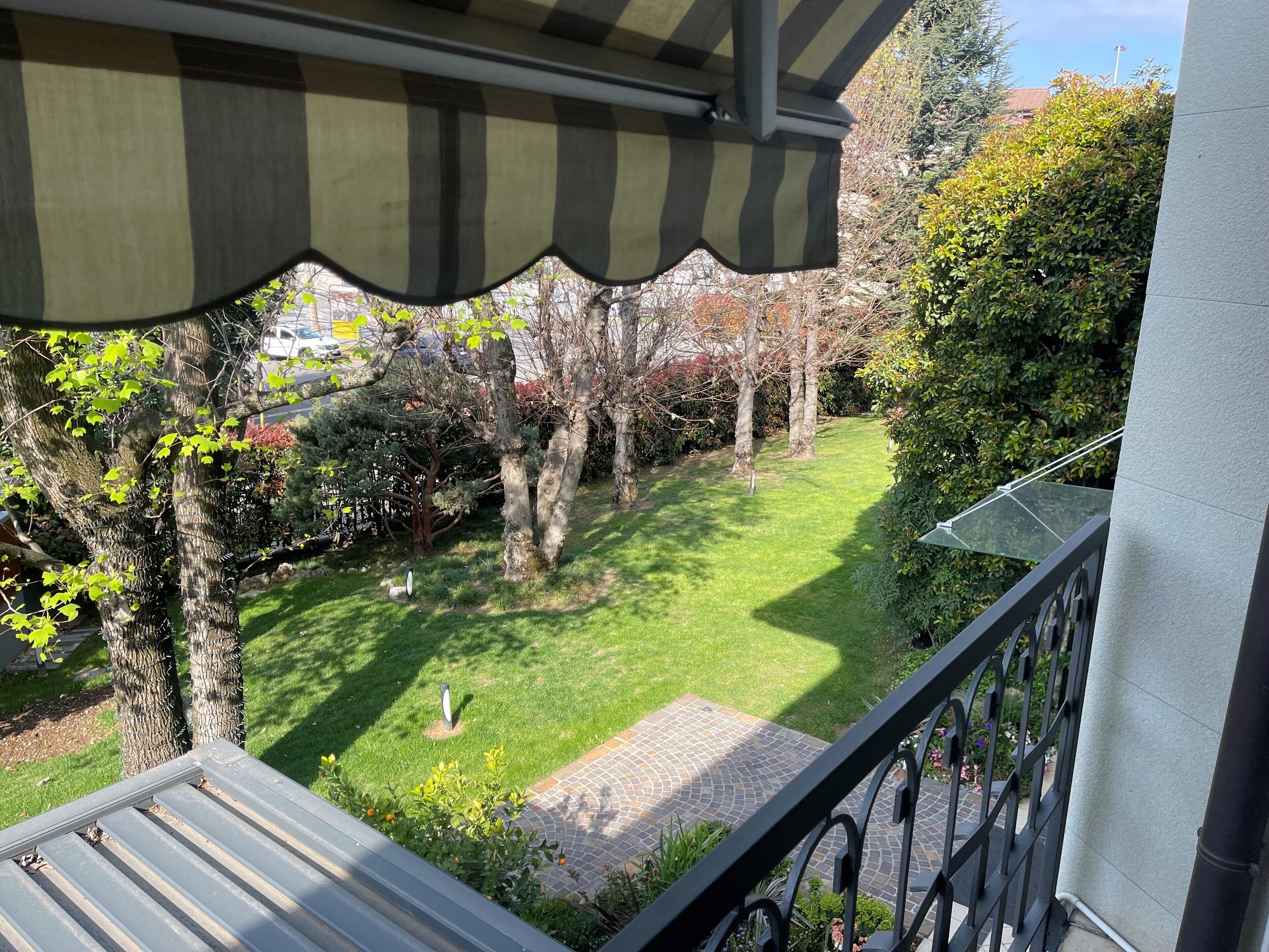 A balcony with a view of a well-maintained garden, including a lawn and various trees.