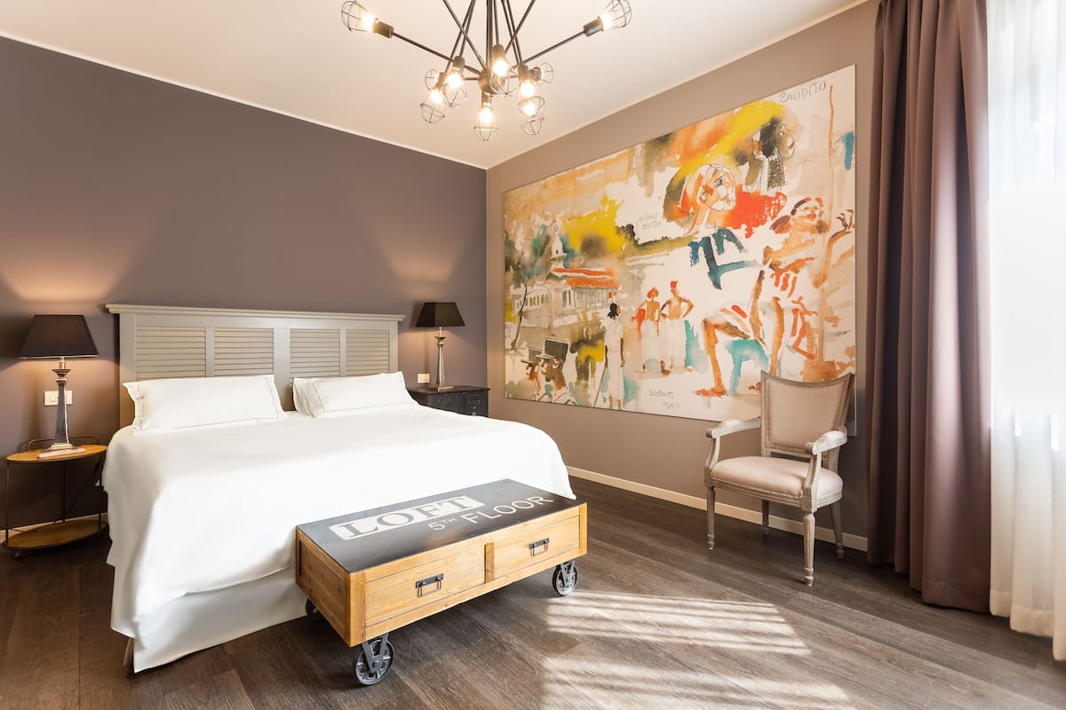 Una habitación de hotel con una cama grande, una mesita de noche de madera, una silla, un tocador, una lámpara y un cuadro abstracto de gran tamaño en la pared.
