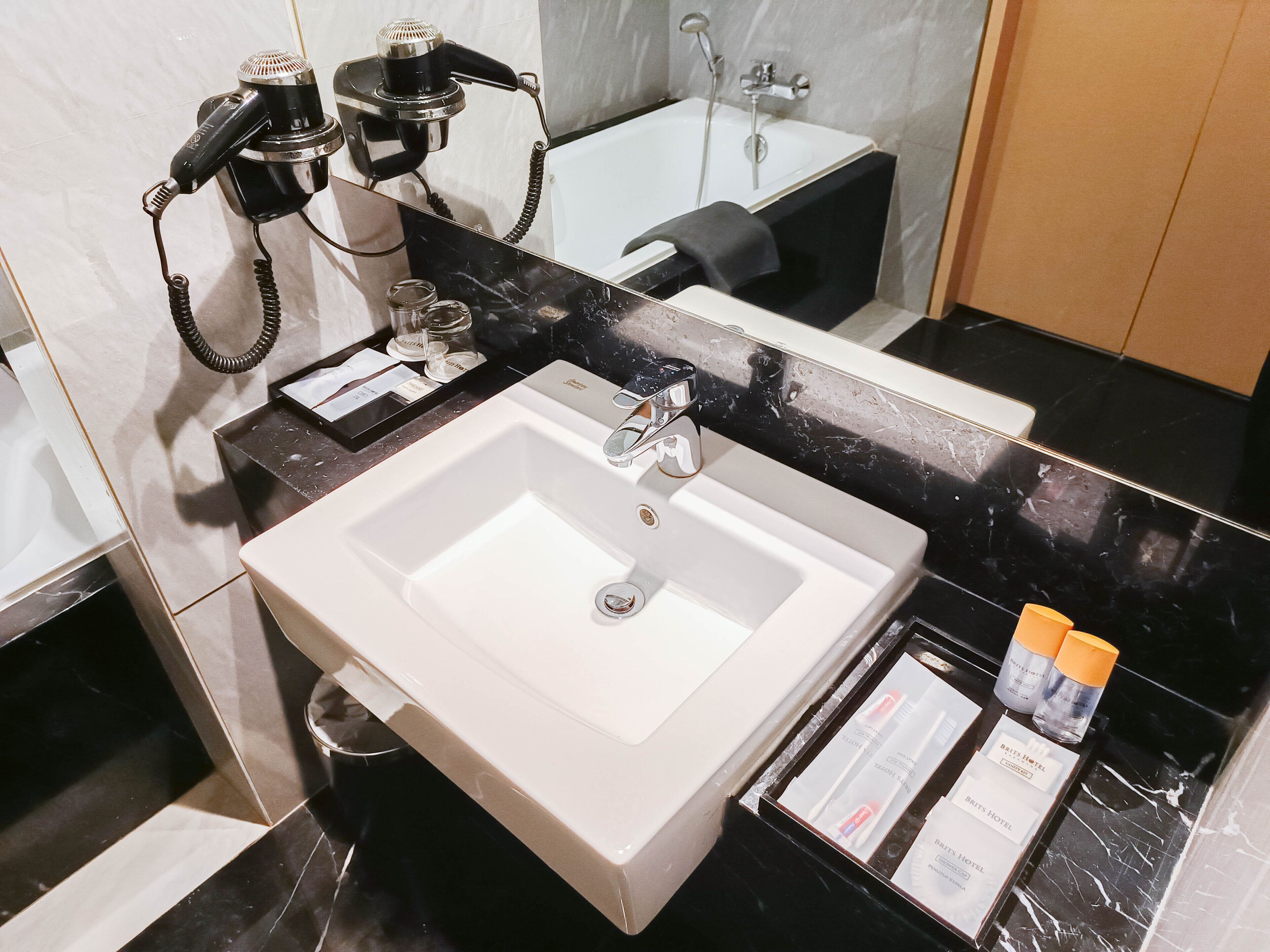 Un bagno con un lavandino bianco, uno specchio e un asciugacapelli. In primo piano, sulla superficie, sono presenti articoli da toilette.