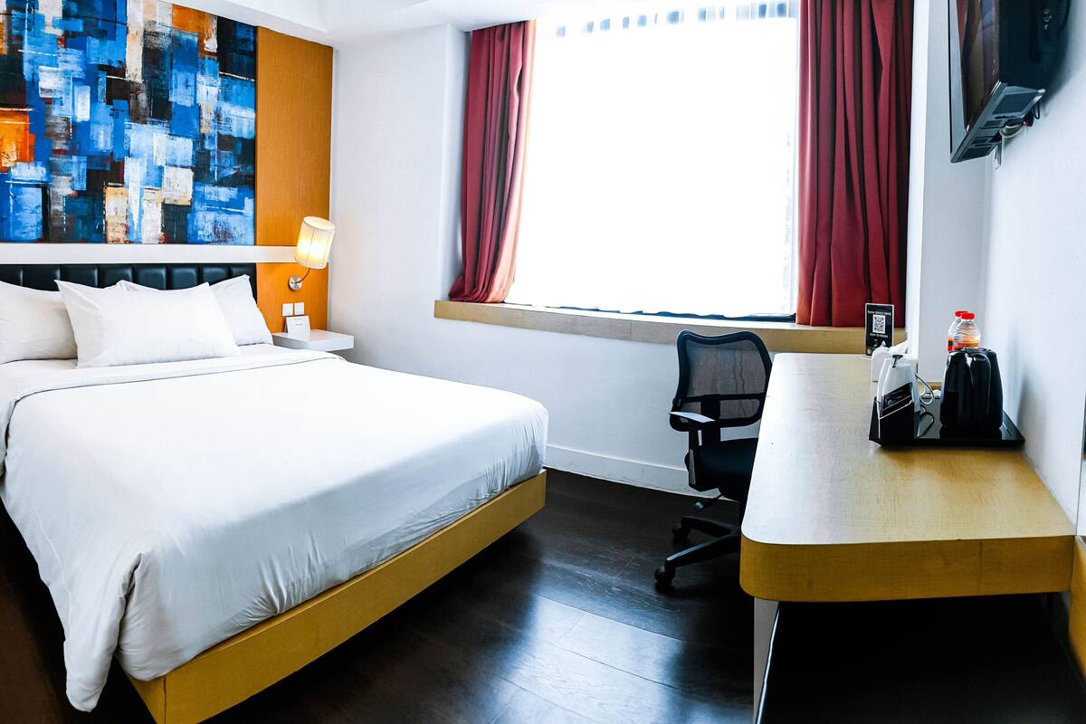 Une chambre d’hôtel avec un grand lit, des tables de chevet avec des lampes, un bureau avec une chaise, une fenêtre avec des rideaux et une peinture abstraite aux couleurs vives accrochée au mur.