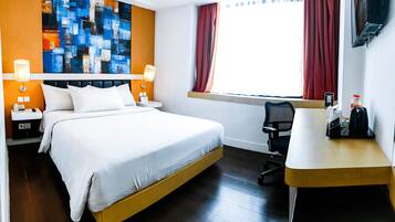 A hotel room with a large bed, bedside tables with lamps, a desk with a chair, a window with curtains, and a colorful abstract painting on the wall.