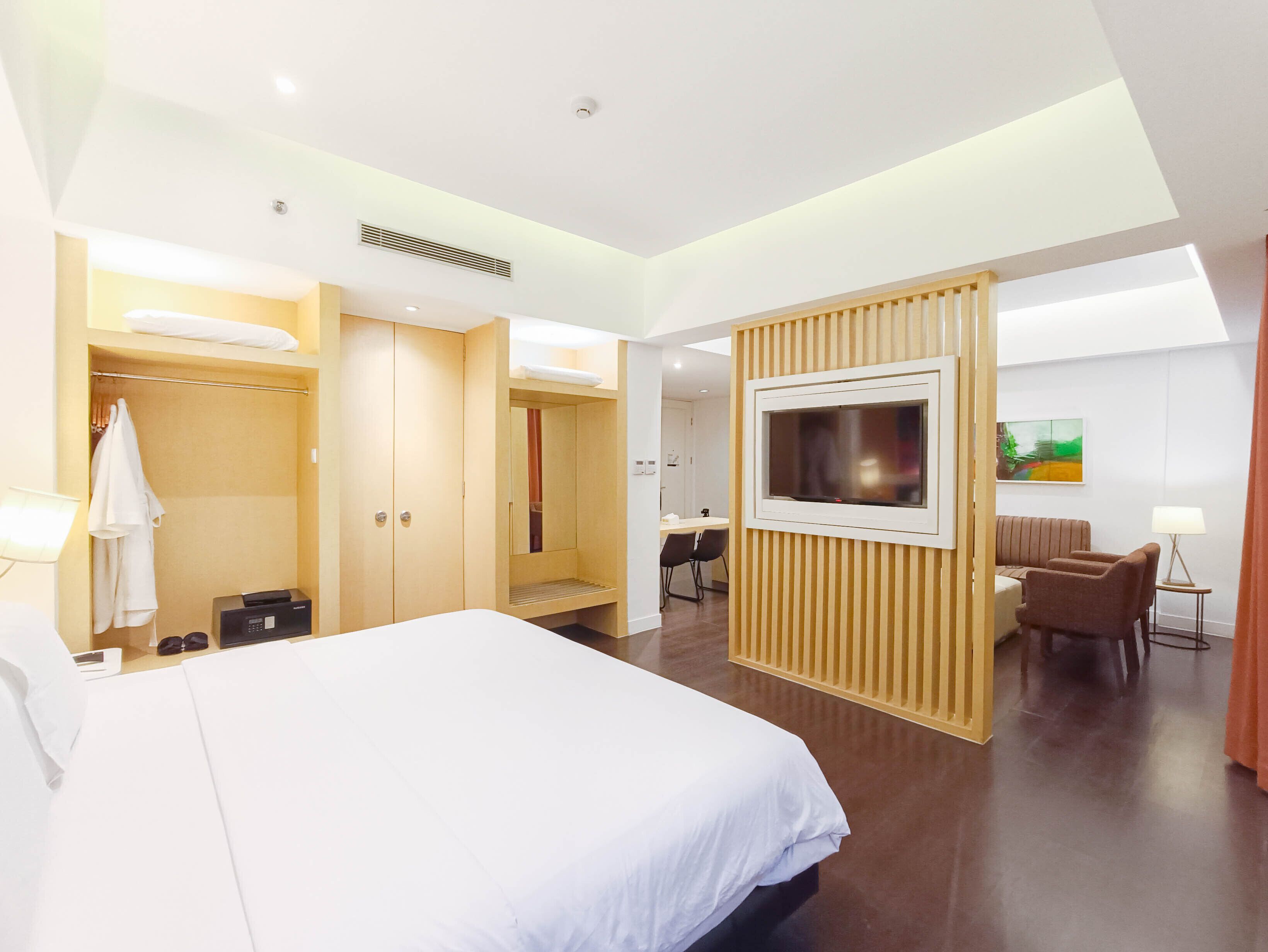 A modern hotel room with a large bed, a desk with a chair, a TV mounted on a wooden panel, and a painting on the wall.