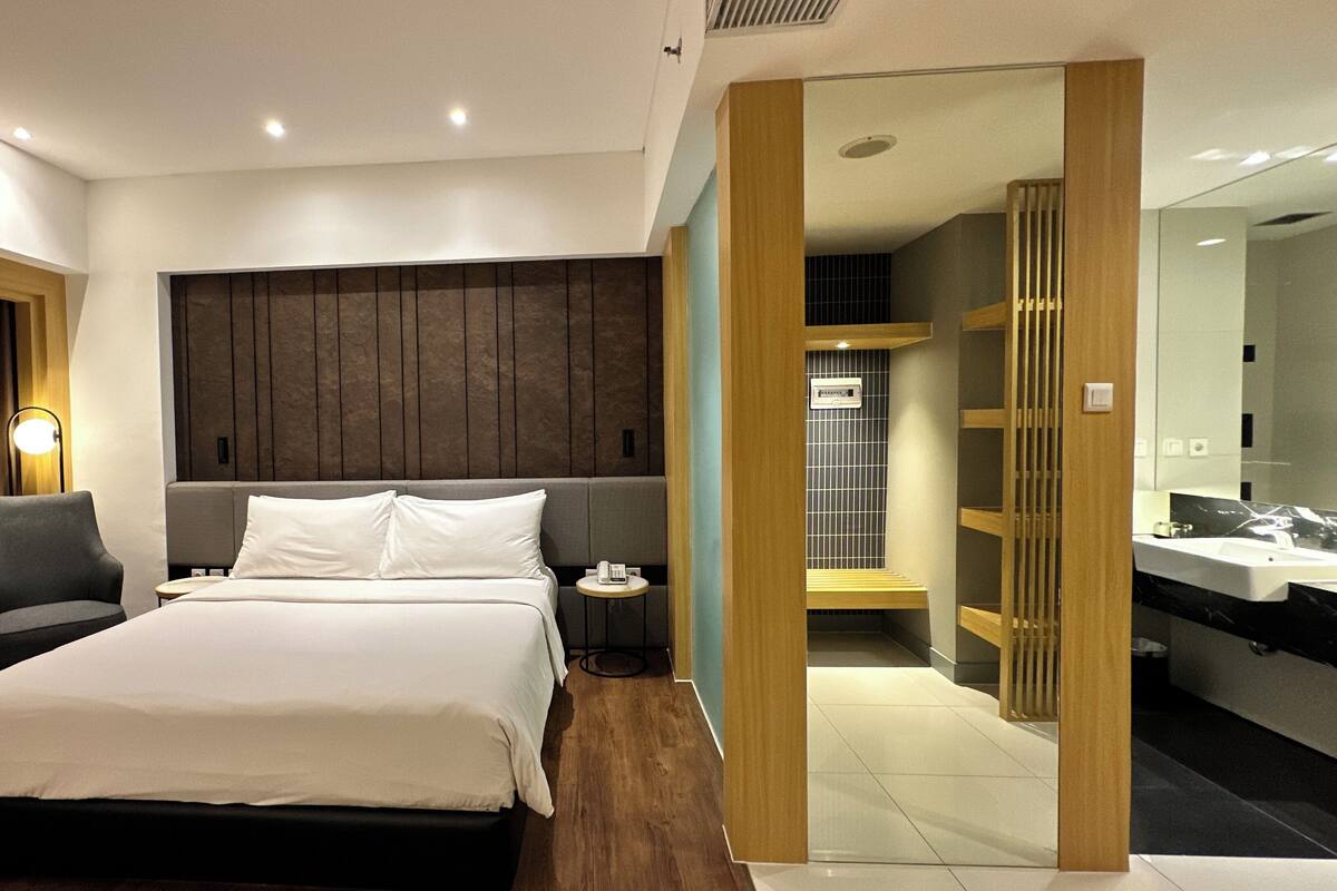 Une chambre d’hôtel moderne avec un grand lit, une table de chevet, une chaise et une petite table ronde. Un mur recouvert de panneaux de bois se trouve derrière le lit, et on aperçoit la salle de bain à travers une porte ouverte.
