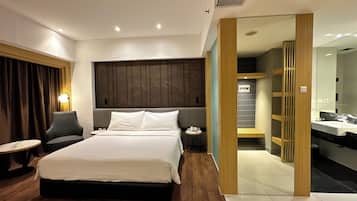 A modern hotel room with a large bed, a bedside table, a chair, and a small round table. There is a wooden panel wall behind the bed and a bathroom visible through an open door.