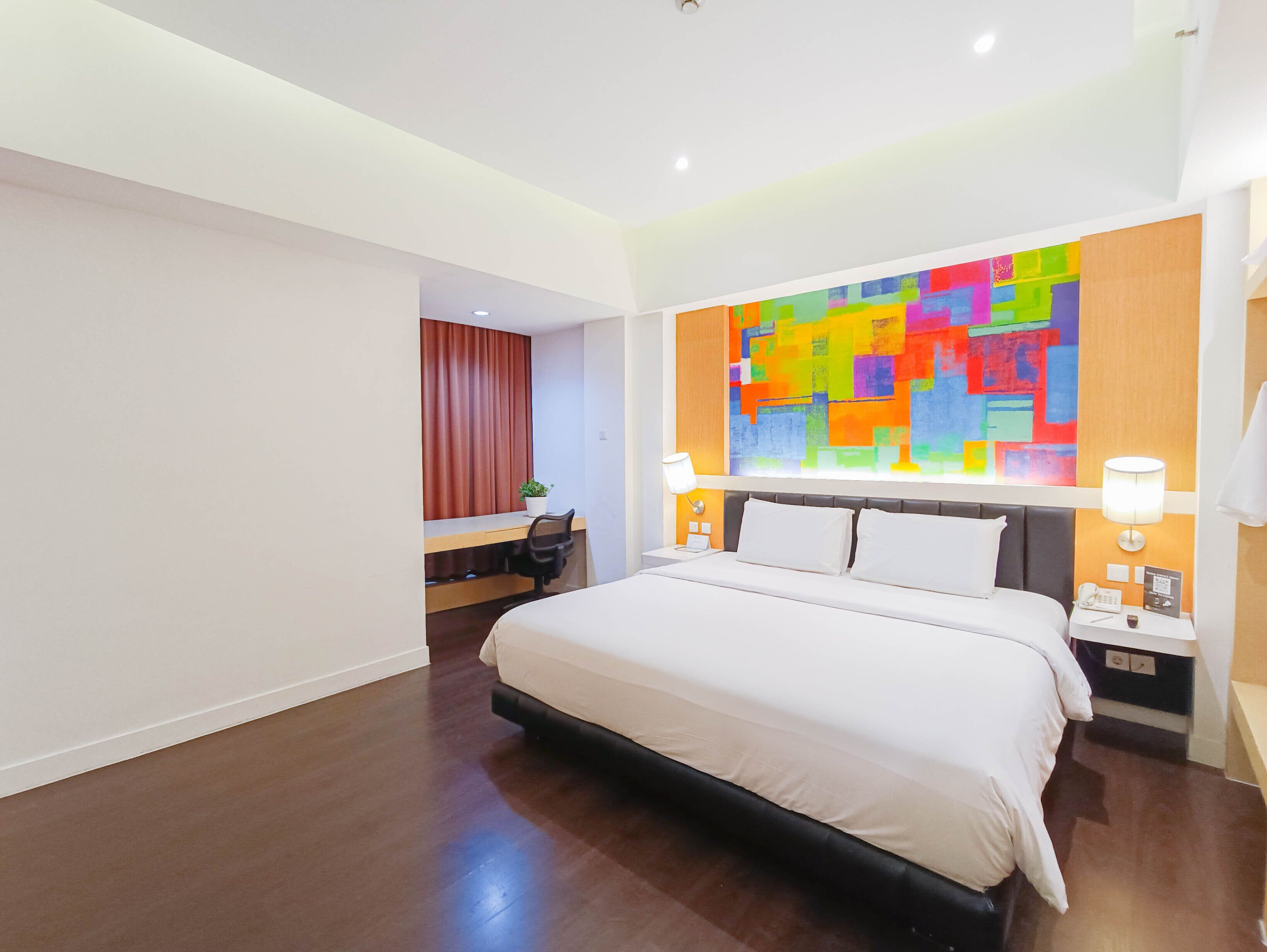 A hotel room with a bed, desk, and chair, a colorful abstract painting on the wall, and a nightstand with a lamp.