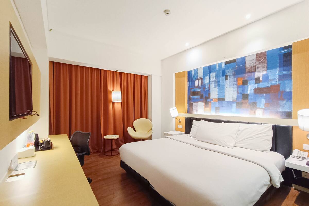 Une chambre d’hôtel comprenant un lit, un bureau, une chaise, une petite table et un mur orné d’un tableau abstrait aux couleurs vives.