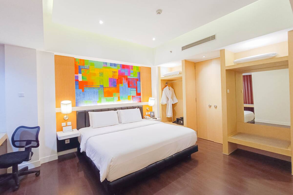 Une chambre d’hôtel avec un grand lit, des tables de chevet, un bureau, une chaise, une peinture abstraite aux couleurs vives, un peignoir et une armoire.