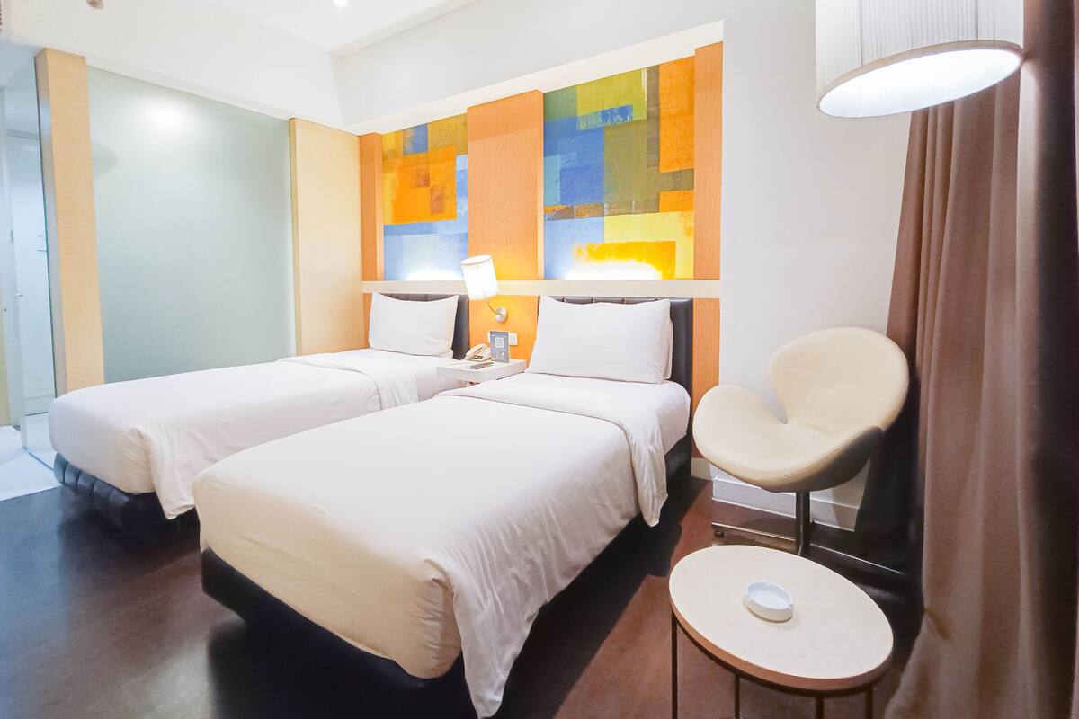 Une chambre d’hôtel avec deux lits, une chaise, une petite table et un tableau abstrait aux couleurs vives accroché au mur.