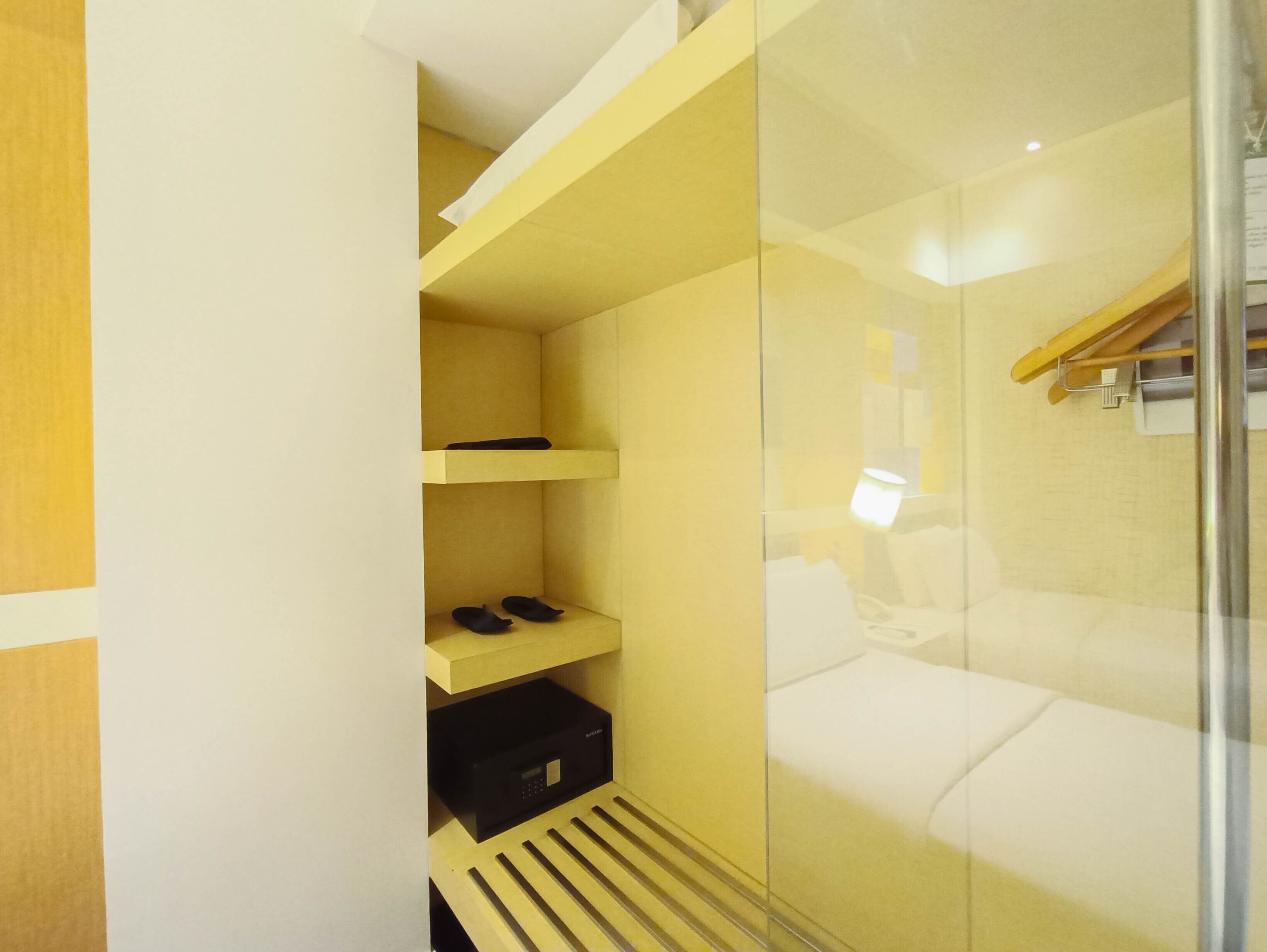 Habitación de hotel con cabina de ducha de vidrio, una cama, un pequeño estante con zapatos y un dispositivo electrónico negro en el suelo.