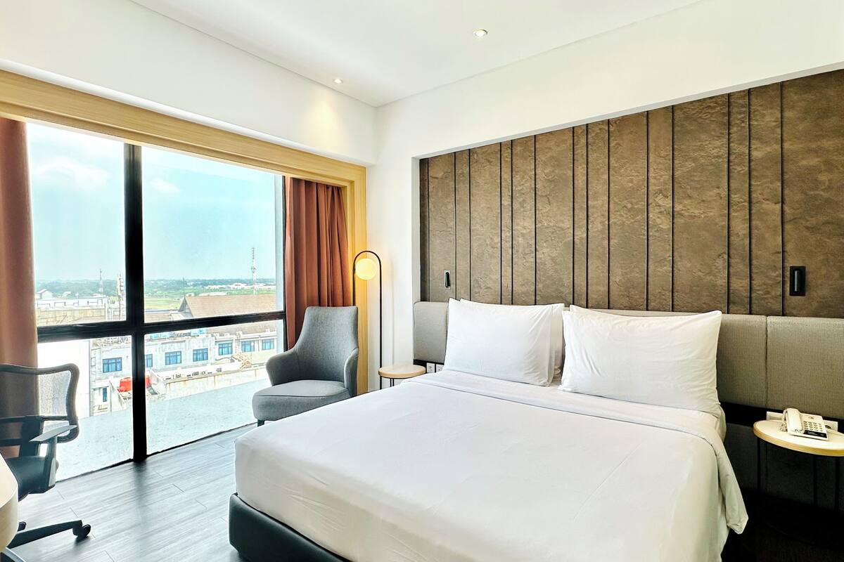 Une chambre d’hôtel dotée d’un grand lit, d’un bureau avec une chaise, d’une petite table avec un téléphone et offrant une vue sur la ville grâce à de grandes fenêtres.