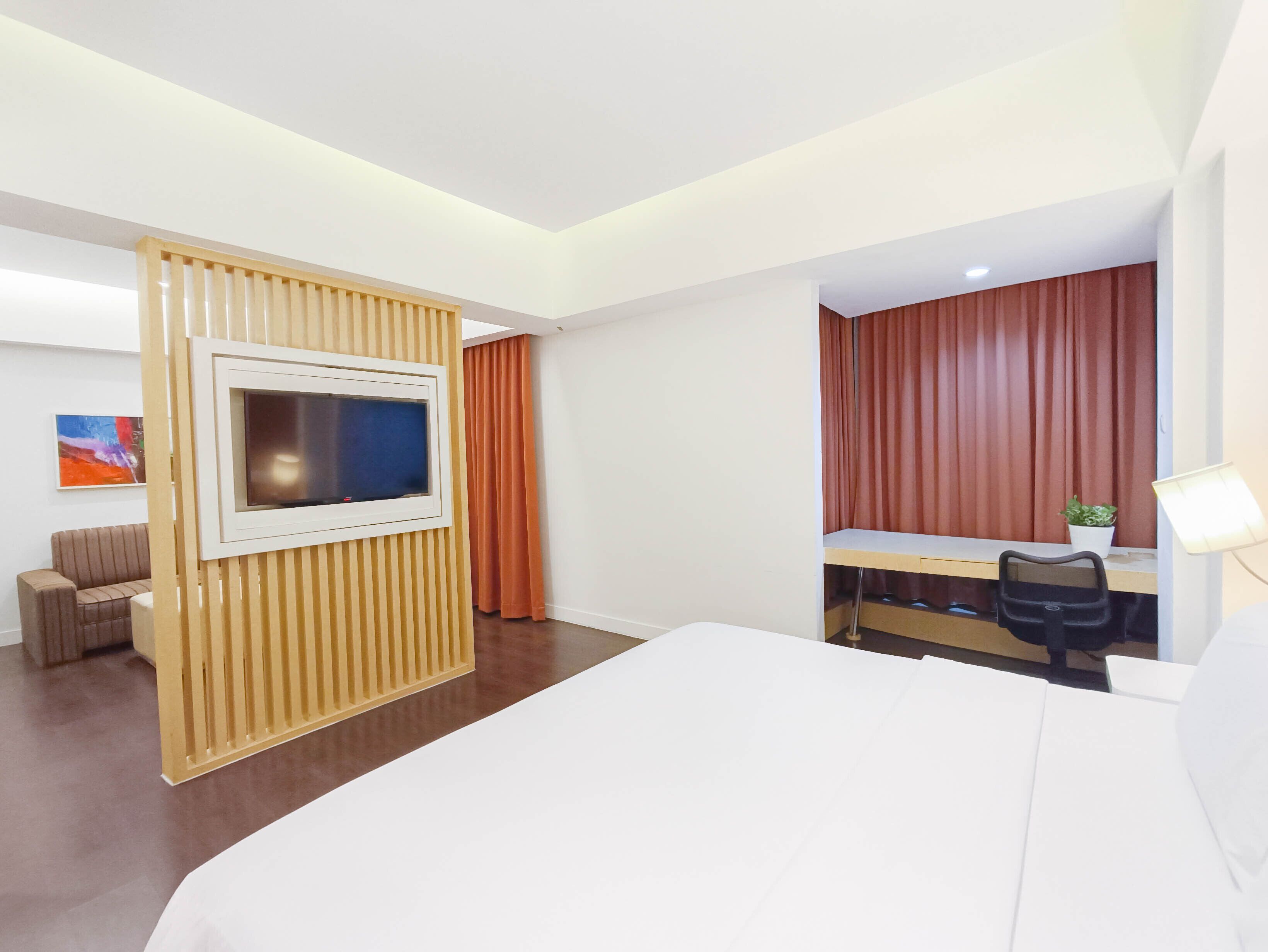 A hotel room with a bed, a desk with a chair, a TV mounted on a wall, a potted plant, and a painting on the wall.