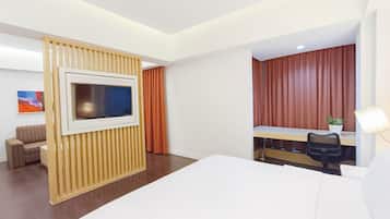 A hotel room with a bed, a desk with a chair, a TV mounted on a wall, a potted plant, and a painting on the wall.