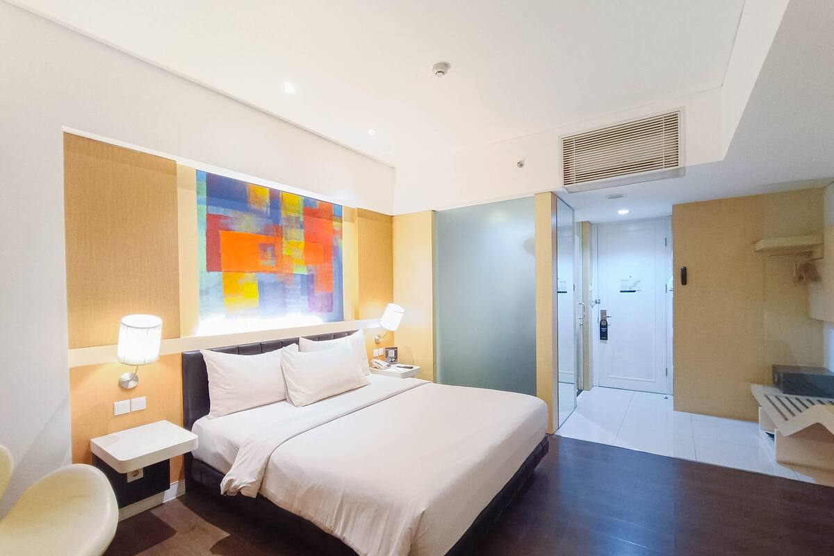 Une chambre d’hôtel moderne dotée d’un grand lit, de tables de chevet, d’un tableau abstrait aux couleurs vives et d’une salle de bain séparée.