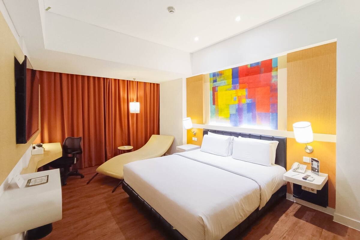Une chambre d’hôtel avec un grand lit, un bureau avec une chaise, une petite table et une peinture abstraite aux couleurs vives accrochée au mur.