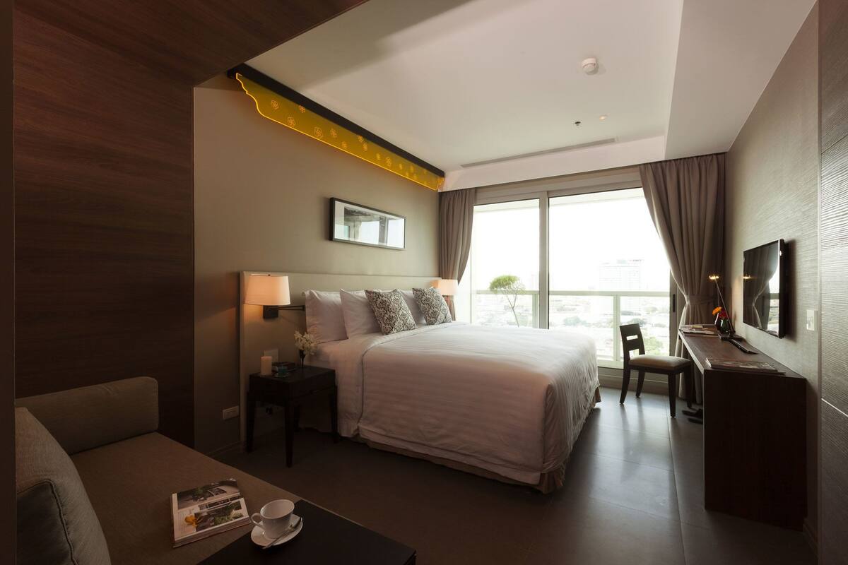 Une chambre d’hôtel avec un grand lit, un bureau avec une télévision, une chaise, une table de chevet avec une lampe et une fenêtre donnant sur la ville.